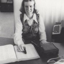 A black and white photo of a young woman using the prototype Optacon machine. She runs a small black probe connected to a larger device over a page in a book.