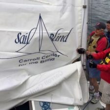 A group of three blind and visually impaired sailors rig a sail on a sailboat that features the SailBlind logo.