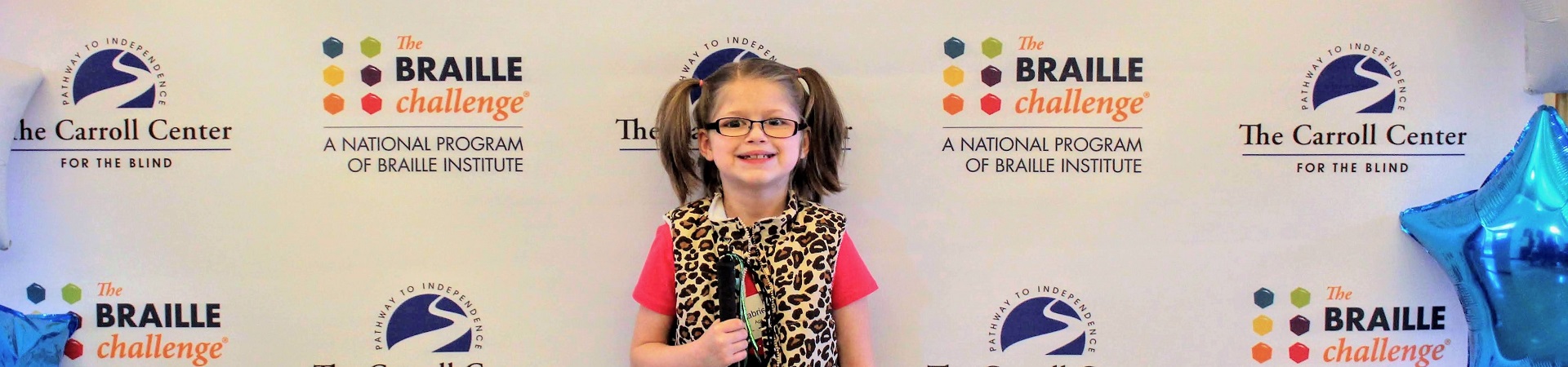 During the New England Regional Braille Challenge, a young visually impaired girl smiles widely standing in front of a "red carpet" backdrop.