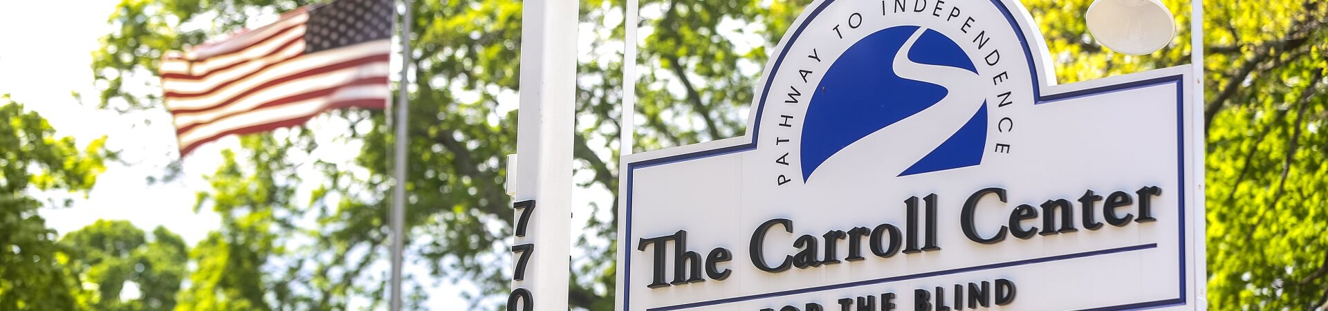 A close-up of the Carroll Center for the Blind sign that hangs at the front of the driveway. An American flag waves in the background.