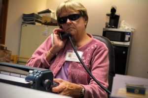 Jan Pecorari, Executive Assistant to the President, talks on a corded phone while typing on the Perkins Brailler on her desk.