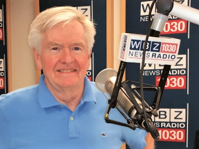 Wearing a blue polo shirt, Dan Rea smiles behind a large microphone with the WBZ 1030 NewsRadio logo on it.