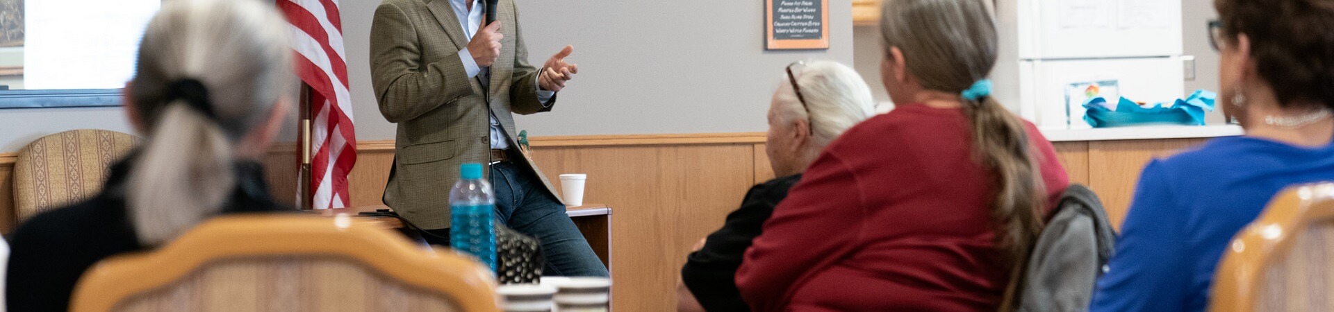 A man holding a microphone presents to a crowd of elderly people at a senior center.