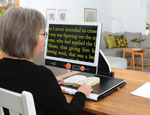 A woman sits down at her desk to read a book using a portable desktop video magnifier.