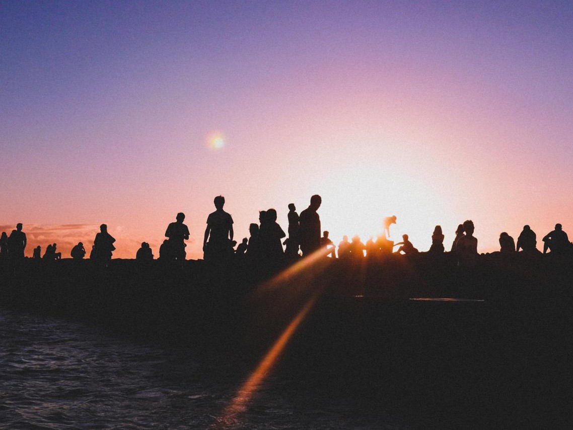 The silhouettes of many people gathered on a beach. The sun shines on the horizon just over their outlines.