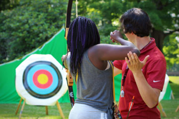 A young female camper in the Carroll Center's summer program takes aim at an archery target with the help of a male instructor.