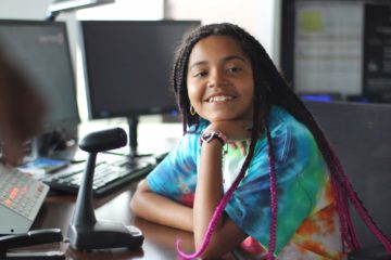 In the United Control Center, a female camper in the Carroll Center's summer program smiles while seated behind a microphone.
