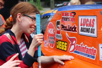 A young woman enrolled in a carroll center summer program feels the car body of the orange dragster.