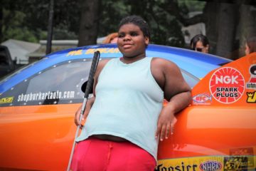 A young woman enrolled in a carroll center summer program leans back and rests on Jay Blake's dragster while grinning.