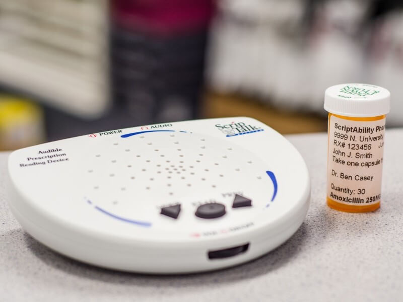 A prescription bottle is placed beside a small, white, battery-operated device called a ScripTalk Station that reads aloud an RFID tag on the bottle.