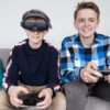 Two young boys sit on a couch and play video games. One boy is wearing an eSight 4 headset.