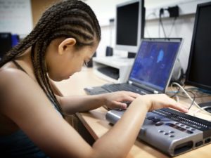 A young woman who is visually impaired uses a refreshable braille display to access the web on her laptop.