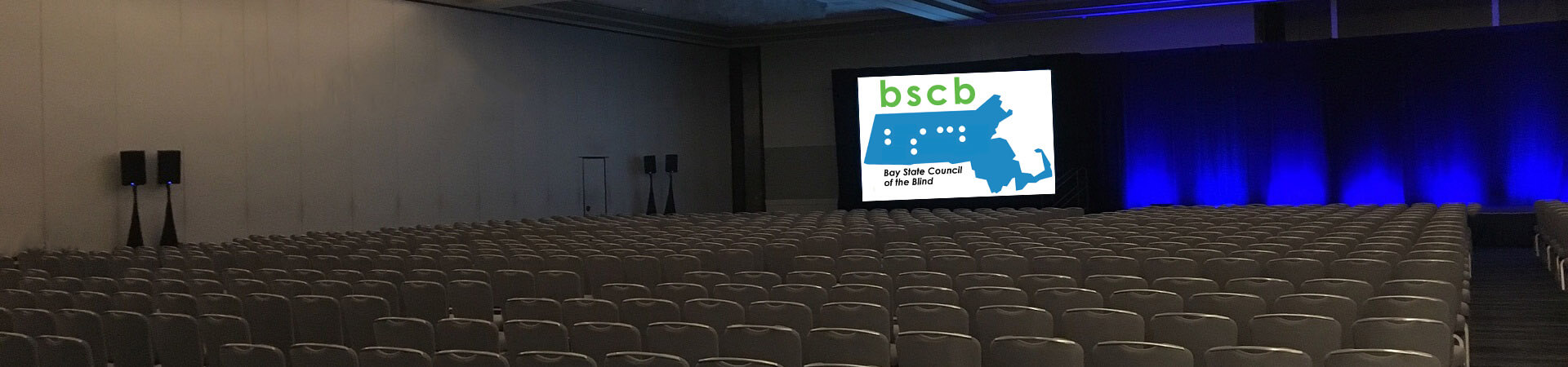 A large auditorium has a BSCB logo projected on the screen by the stage.