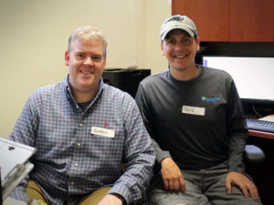 Two male volunteers smile together as they take a break from administrative work on computers at the Carroll Center for the Blind.