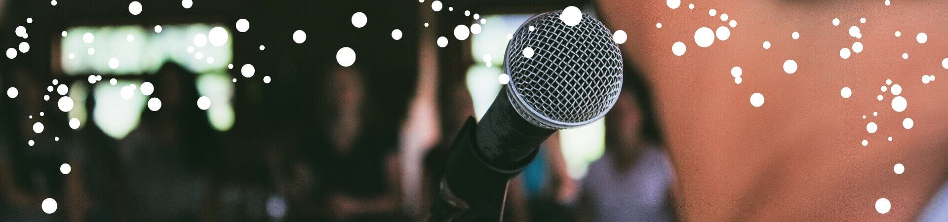 A close-up of a microphone from behind a performer's head. A blurry audience can barely be made out in the background.