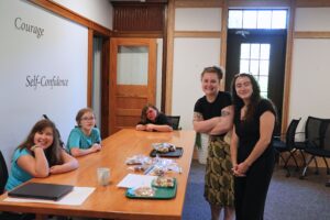 2 smiling coworkers stand beside a table sitting 3 summer program students as they have their bake sale.