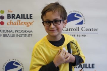 Apprentice group winner, Benjamin Silva, wears a yellow braille challenge shirt as he poses in front of a braille challenge banner.