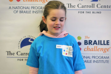 Freshman group winner, Hannah Gevers, wears a blue braille challenge shirt and poses in front of a braille challenge banner.
