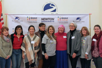A group of 9 staff and volunteers pose in front of a banner at the Braille challenge.