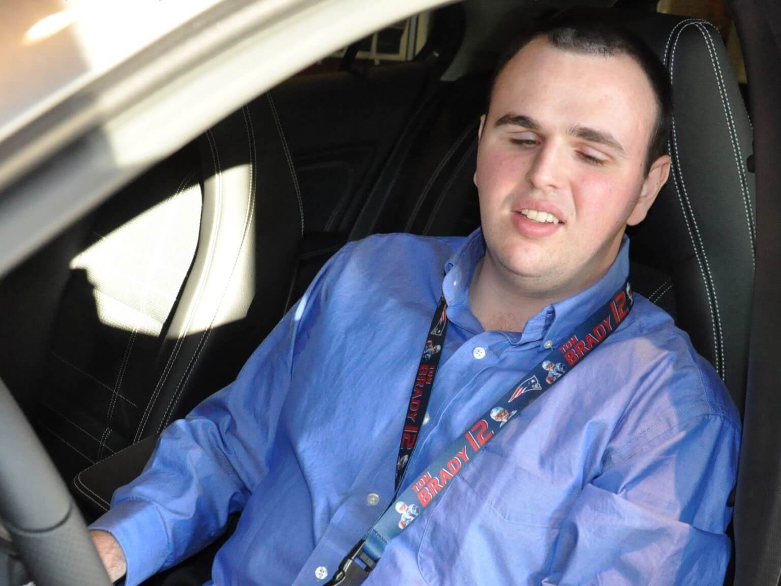 Adam Roberge, wearing a blue button down shirt with a Patriots lanyard around his neck, smiles as he sits in the driver's seat of a Mercedes-Benz on his first day of work at the dealership.