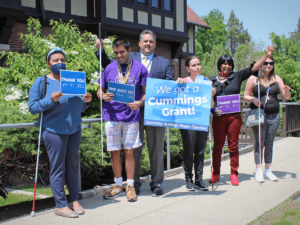 Gregory J. Donnelly, President and CEO of the Carroll Center for the Blind, celebrates the Cummings Foundation Grant by holding up signs with five Carroll Center clients outdoors. The signs read, "We Got a Cummings Grant!"