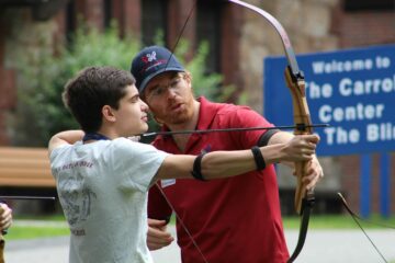 An instructor works with a youth in transition student during archery.