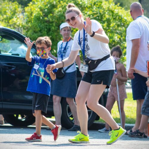 An instructor and Carroll Kids student hold hands and wave at the camera as they walk across a parking lot.