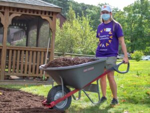 A volunteer from Point 32 Health wearing a purple company shirt empties a wheelbarrow filled with dirt in front of a gazebo at the Carroll Center for the Blind.