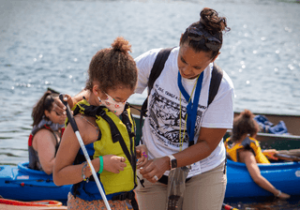Carroll Center volunteer helping a young summer student zip on her life vest before going kayaking.