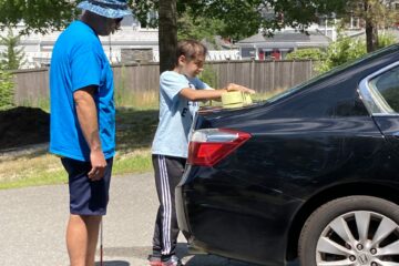 Blue shirted supervisor with a light blue shirted young boy washing a black car
