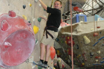 A boy with a black shirt in the middle of a climbing wall looking back at the camera