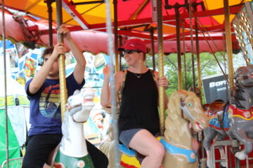 Smiling supervisor next to a dark blue wearing teen both on a horse on a carousel