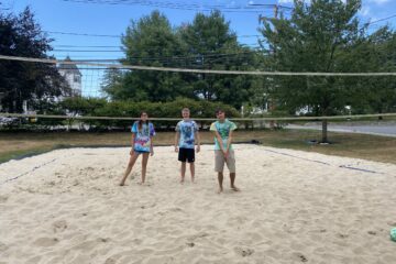 Three students, a guy in a green shirt, a guy in a blue shirt, and girl in a blue shirt, are on sand to play volleyball.