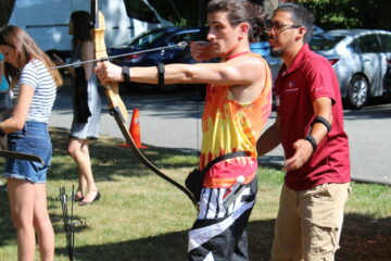 A boy wearing a tie dye shirt shooting a bow and arrow with adult supervision