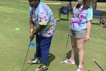 Two teen girls in tie dye shirts taking turns hitting the golf balls into a hole.