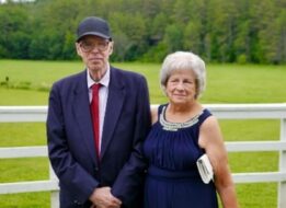 Peter and Deborah Coogan pose together in front of a white fence with a forest behind it.