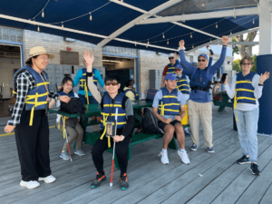 Group of SailBlind sailors, instructors, and volunteers standing near and sitting at a picnic table on the dock raising their hands and waving in excitement and smiling.