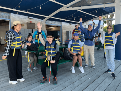 Group of SailBlind sailors and instructors standing near and sitting at a picnic table on the dock raising their hands and waving in excitement and smiling.