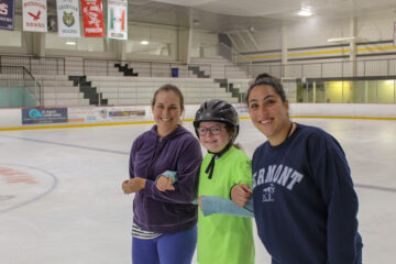 2 smiling instructors linking arms with a Carroll Center student girl on the ice skating rink