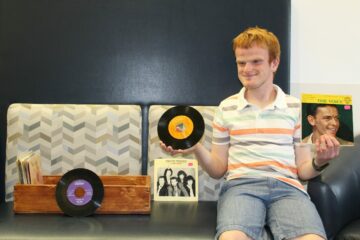 a student sitting on a bench and holding records next to a wooden record box he made in a sensory arts class