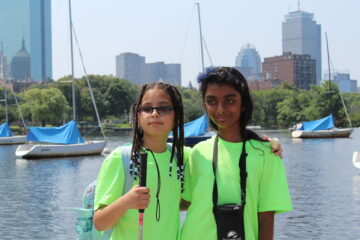 2 girls smiling on the Charles River Esplanade with the Boston skyline in the background