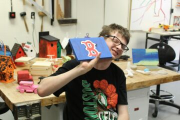 a smiling student smiling holding a painting of a letter "B" from the Boston Red Sox logo which he made in a sensory arts class