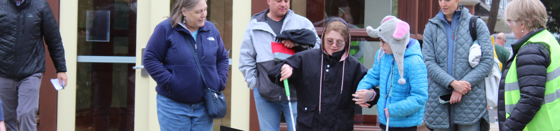 The image shows two young individuals holding white canes at the Cane Quest event last year. Surrounding them are a few adults dressed warmly, one of whom is wearing a reflective safety vest.