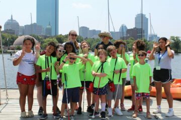 10 students and 4 instructors making silly faces for a group photo on the Charles River Esplanade with the Boston skyline in the background