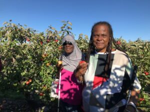 Two Carroll Center alumni are standing in front of an apple orchard on a sunny day. The woman on the left is wearing a gray headscarf, sunglasses, and a pink top, and is holding a white cane in her left hand while holding an apple in her right. The woman on the right has long braided hair, is wearing a multicolored geometric-patterned top, and is also holding an apple. Both women are smiling, and the orchard behind them is filled with trees bearing red apples. The sky is clear and blue.