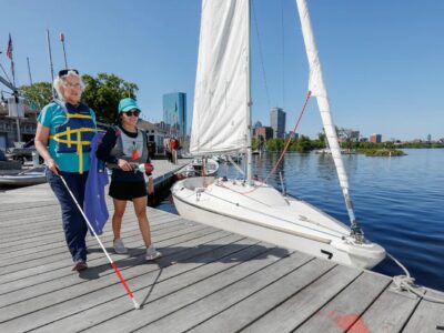 A Sailblind sailor is walking on the dock with a white cane and a sighted guide