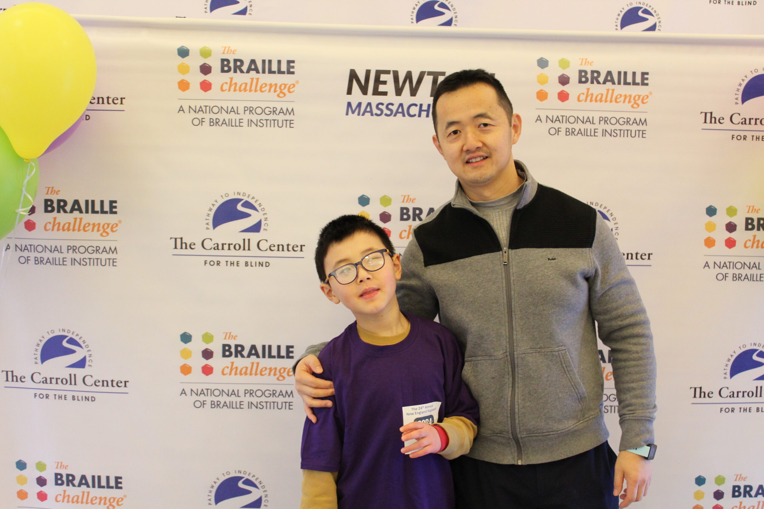 Braille Challenge participant and his father smiling for a photo in front of the Carroll Center Braille Challenge backdrop