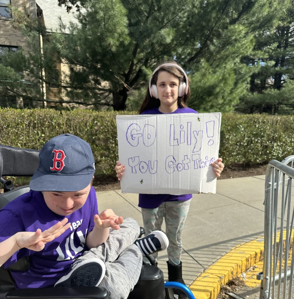Lily's student standing and posing for a picture with a sign that says "Go Lily! You got this" with another student smiling in the background