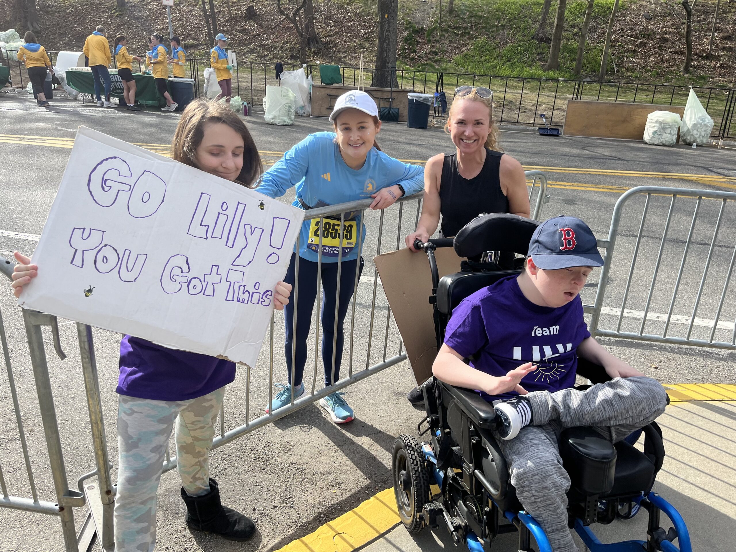 Lily standing next to two of her students and one parent. One of the students is sitting in a wheelchair and the other one is standing and holding a sign that says "Go Lily! You got this"