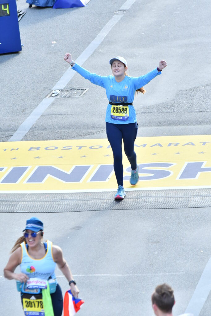 Lily McDonagh smiling at the Boston Marathon finish line, with her hands raised into the air.